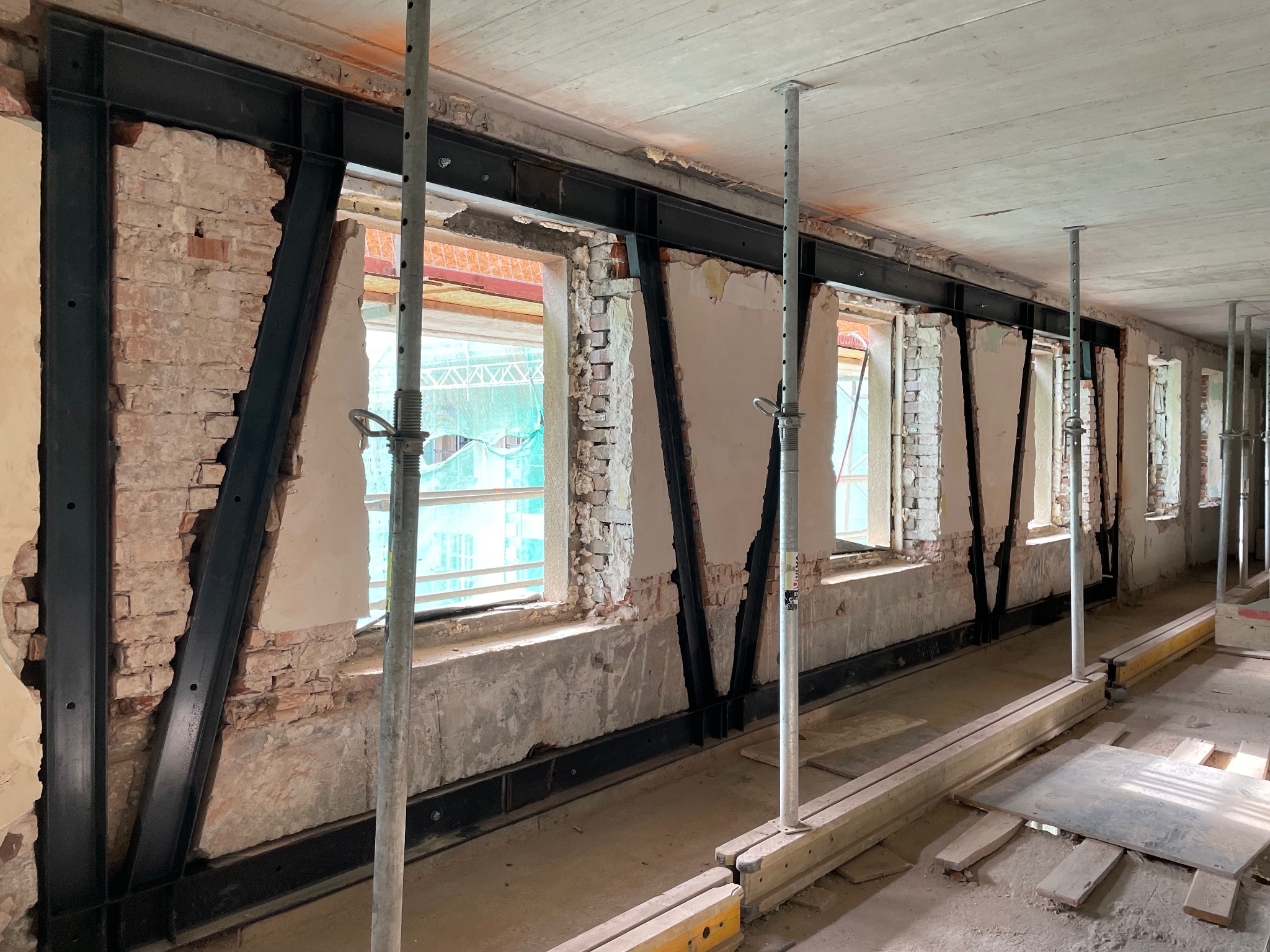 Wnętrze w trakcie remontu: wzmocnione stalowymi belkami okna w surowym murze, widoczne rusztowania i elementy konstrukcyjne. Prace budowlane w starym budynku.