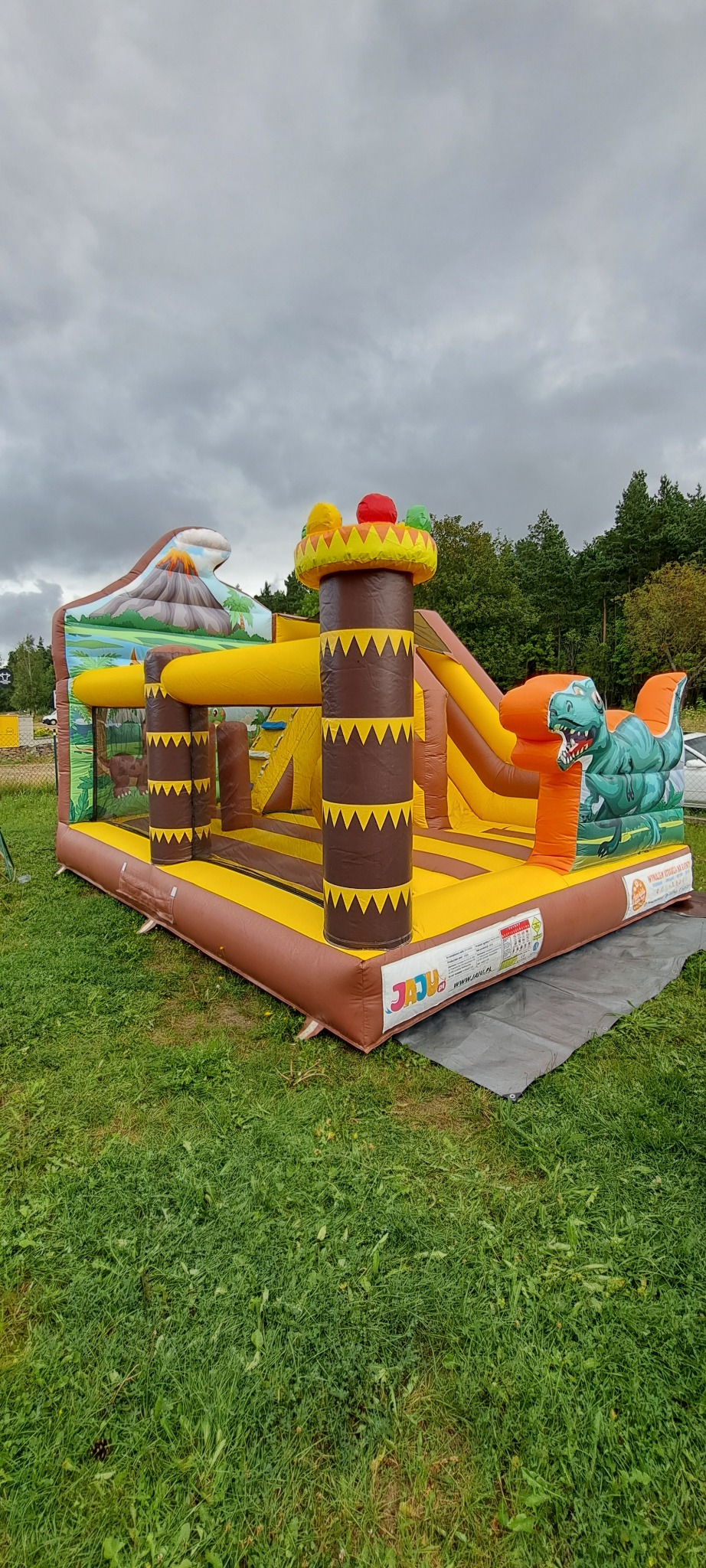 Duży, kolorowy dmuchany zamek z motywem dinozaurów i wulkanu, ustawiony na trawie pod pochmurnym niebem. Widoczne elementy zjeżdżalni i kolumny.