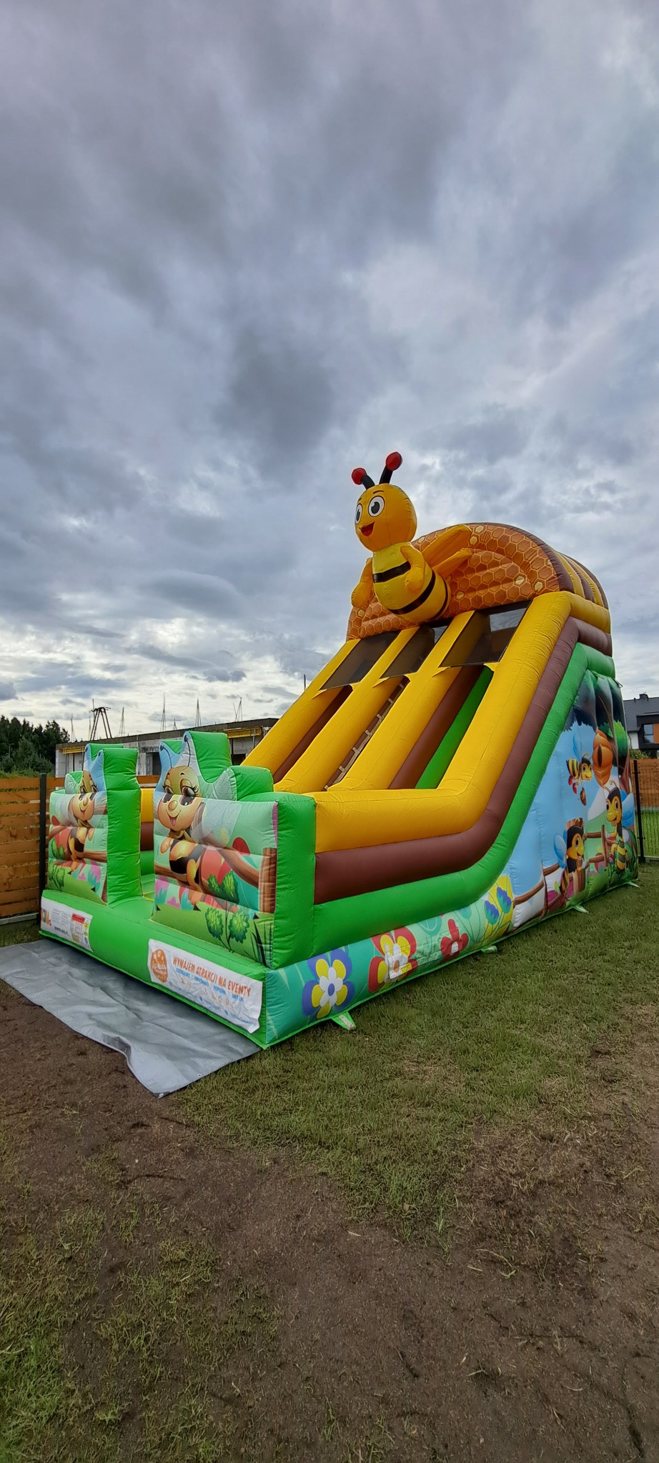 Kolorowa, dmuchana zjeżdżalnia z motywem pszczoły na trawie, pod pochmurnym niebem. Zjeżdżalnia ma żółte pasy i zielone boki z grafikami pszczół i kwiatów.