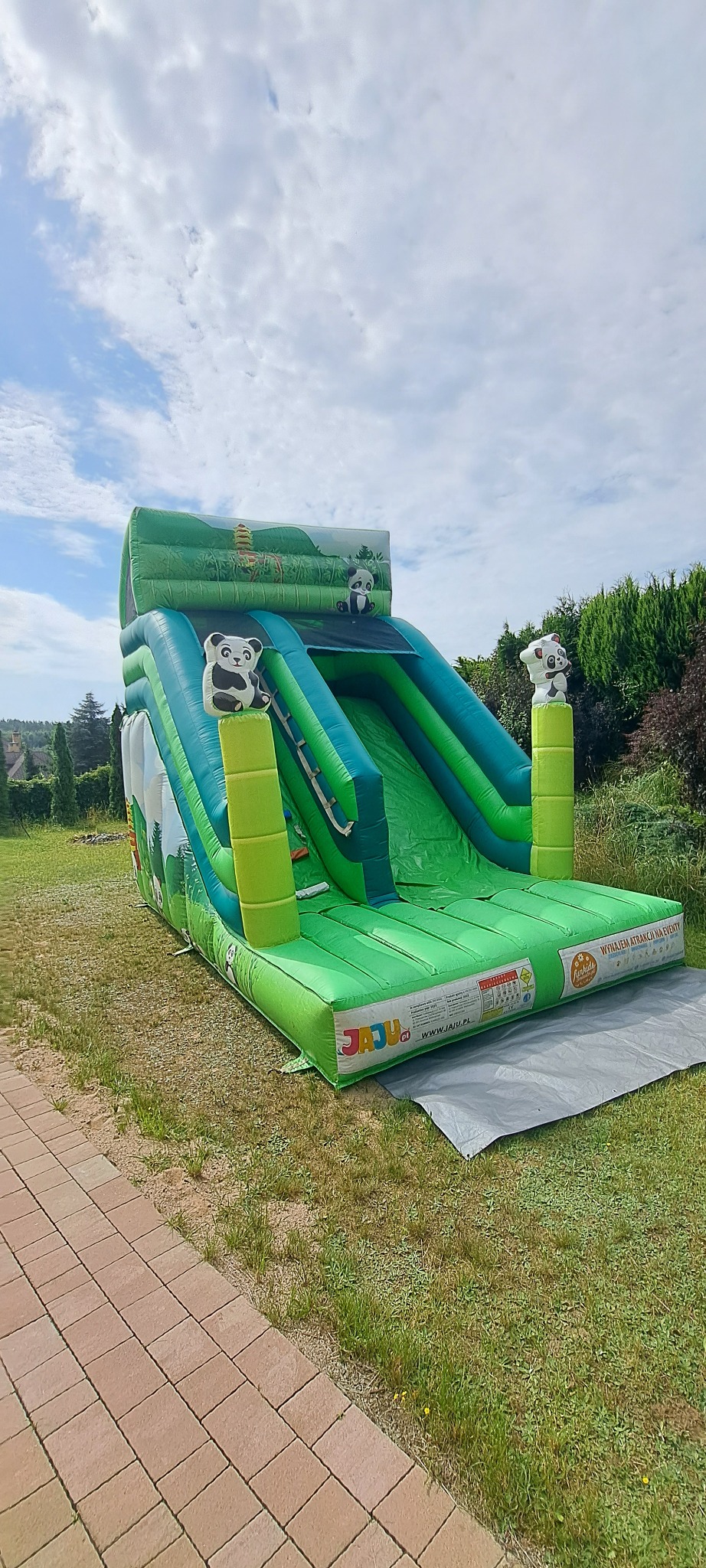 Zielono-niebieska dmuchana zjeżdżalnia z motywem pandy na trawie, pod błękitnym niebem. Widoczne logo firmy na boku. Zjeżdżalnia zabezpieczona szarą matą.