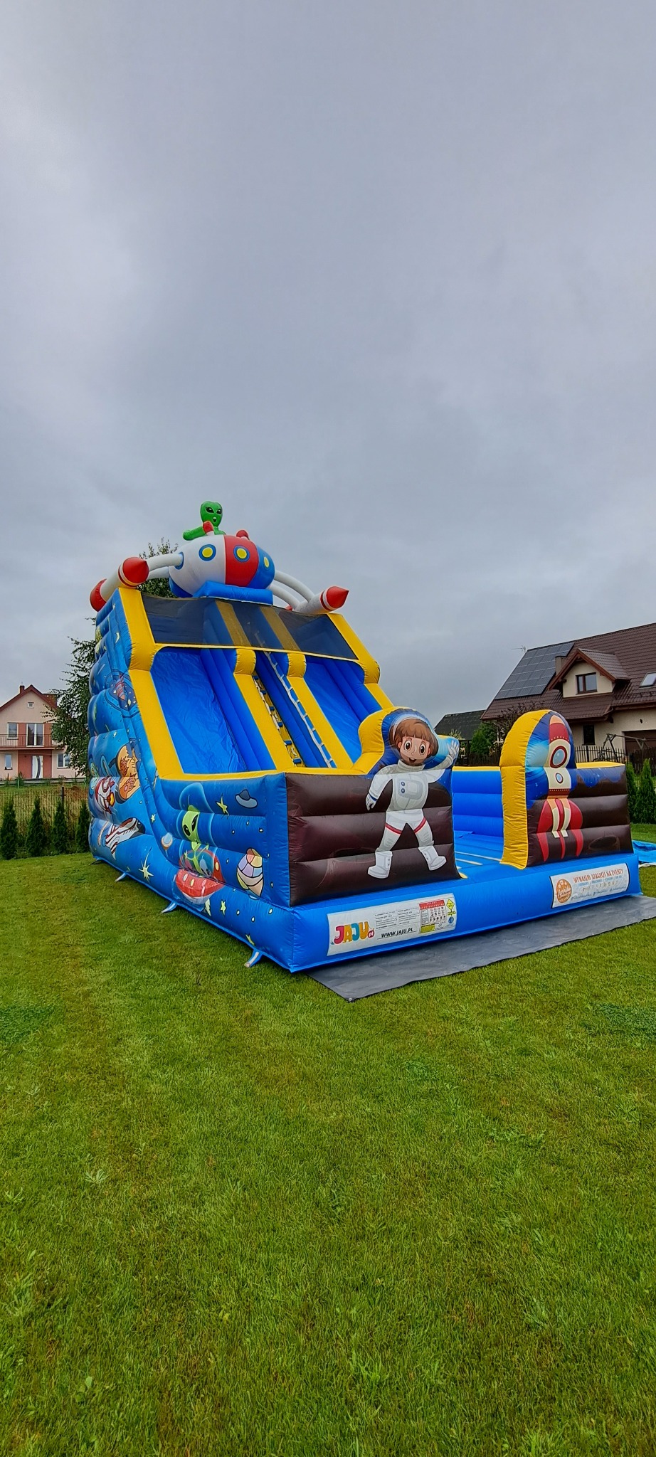 Kolorowy dmuchany zamek w kosmicznym stylu, z motywem astronautów i rakiet, ustawiony na trawniku przed domem. Widoczne zjeżdżalnie i elementy dekoracyjne.
