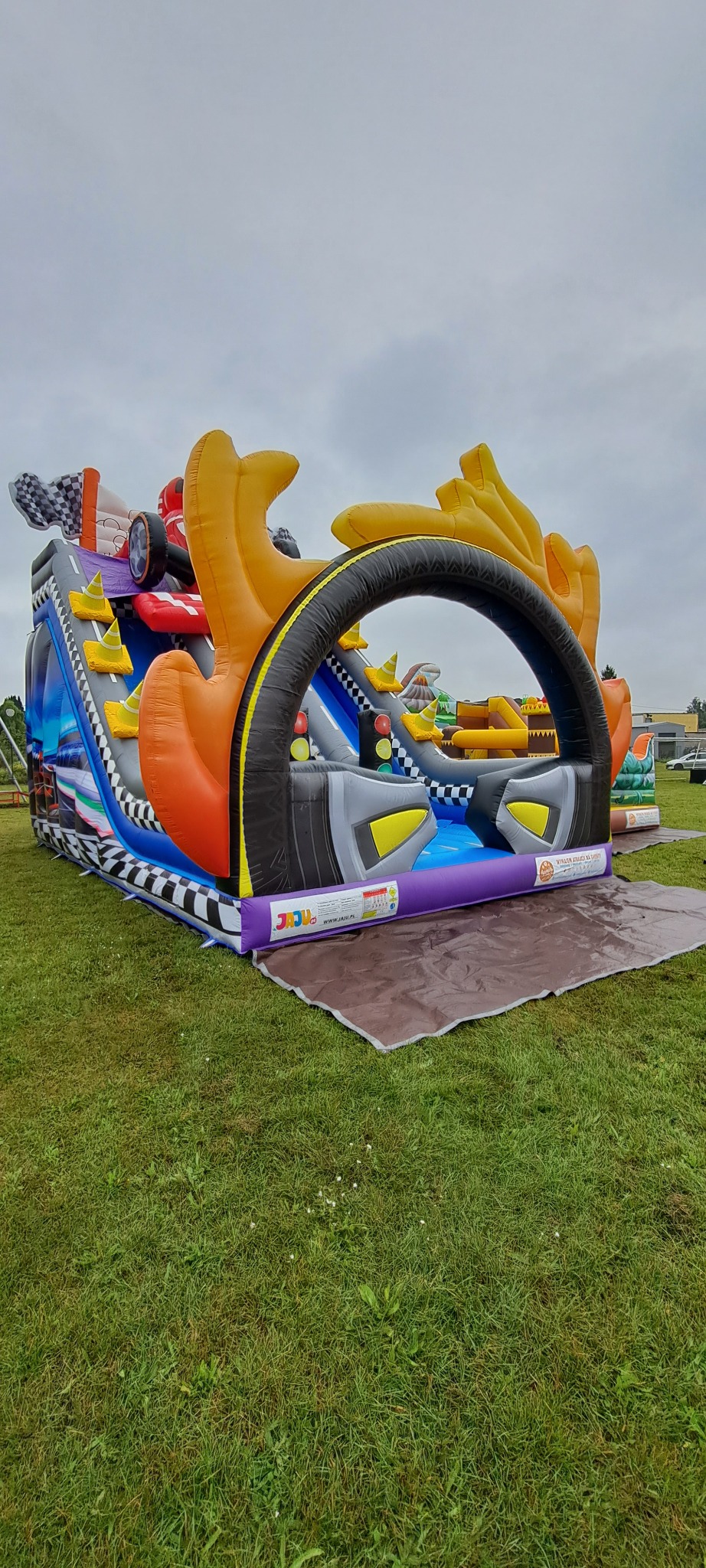 Kolorowy dmuchany zamek w kształcie toru wyścigowego z przeszkodami, na zielonej trawie, pod pochmurnym niebem. Widoczny fragment maty zabezpieczającej.