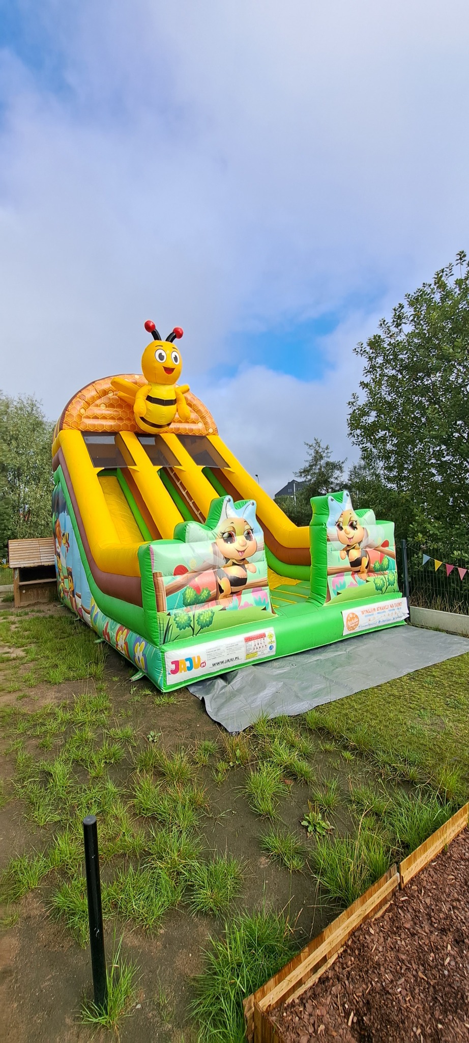 Kolorowa, dmuchana zjeżdżalnia z motywem pszczół, ustawiona na trawie. Widoczne elementy dekoracyjne i drabinka wejściowa. Idealna atrakcja na imprezy plenerowe dla dzieci.