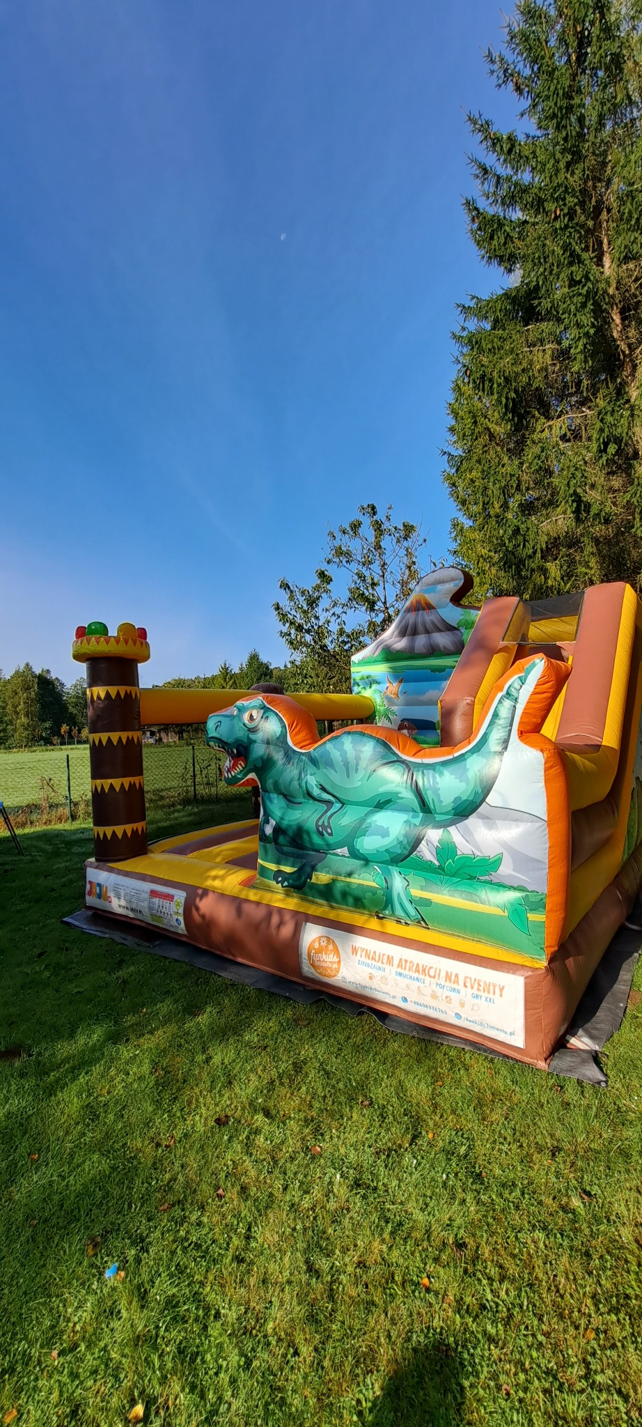 Dmuchany zamek z motywem dinozaura na trawie w słoneczny dzień. Atrakcja eventowa z elementami wulkanu i wieży. Widoczne logo firmy oferującej wynajem.