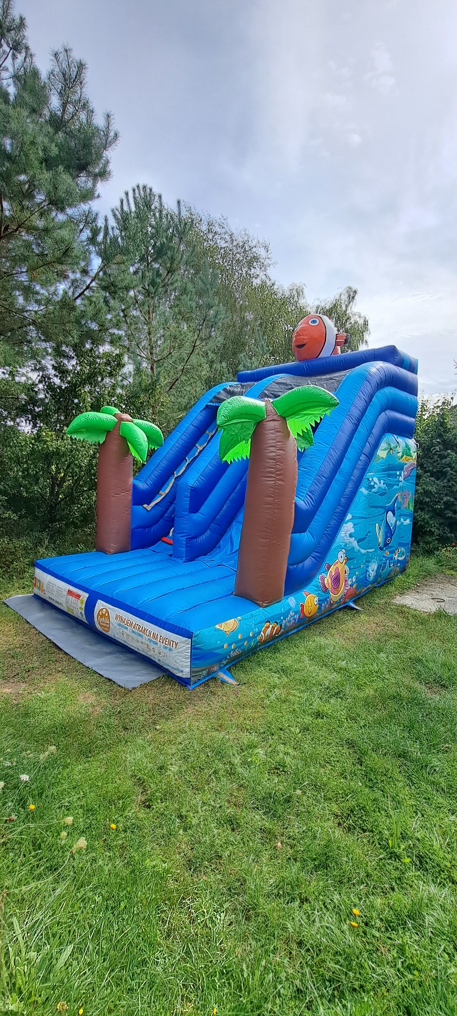 Niebieska, dmuchana zjeżdżalnia z motywem ryb i palm, zwieńczona postacią ryby Nemo, ustawiona na trawniku. Widoczne drzewa w tle.