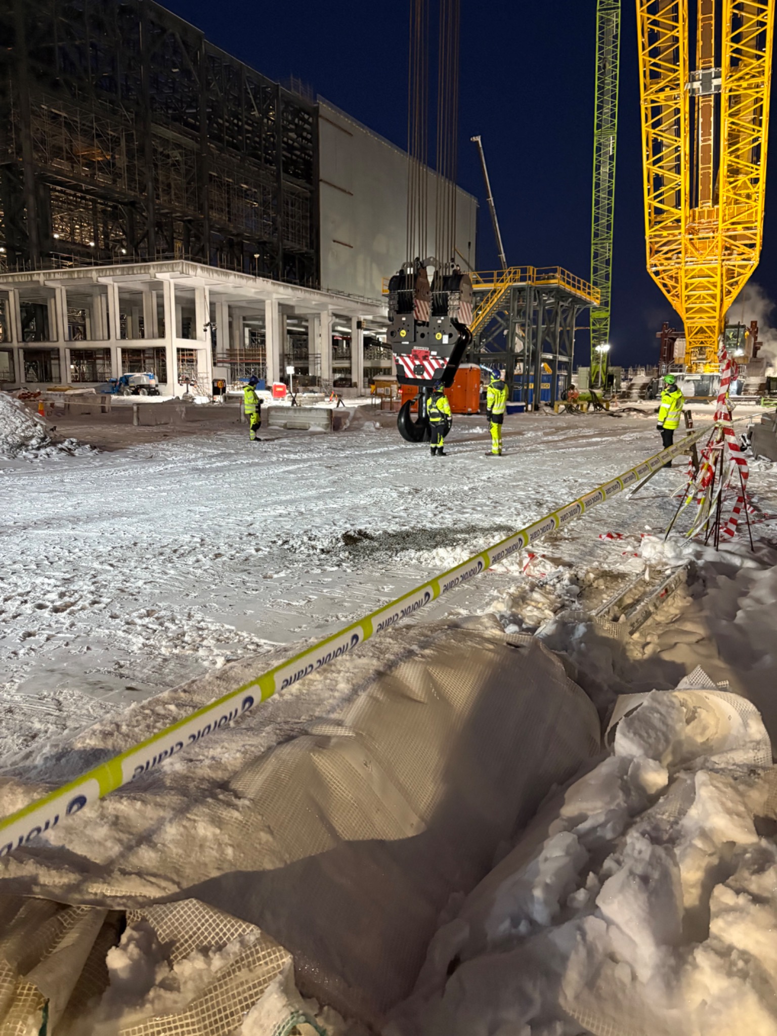 Nocna sceneria budowy: pracownicy w odblaskowych strojach, żurawie i konstrukcja budynku w tle, teren oznaczony taśmą ostrzegawczą na śniegu.