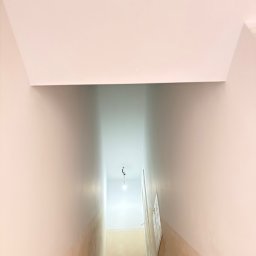 Kacper Starobrzyński - Klatka schodowa w trakcie malowania: górna część ścian i sufit pomalowane na biało, dolna część ścian w stanie surowym, widoczne schody i wiadro z farbą.