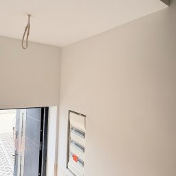 Kacper Starobrzyński - Świeżo pomalowane, jasne wnętrze z widoczną skrzynką elektryczną i wystającym kablem oświetleniowym. Drzwi wejściowe z widoczną kostką brukową na zewnątrz.