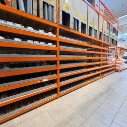 Kacper Starobrzyński - Pomarańczowy regał magazynowy z półkami, na których leżą kartony z izolacją termiczną. Widok z dołu, perspektywa zbieżna, jasne oświetlenie.