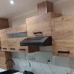 Kacper Starobrzyński - Nowoczesna zabudowa kuchenna z drewnopodobnymi frontami i czarnymi uchwytami. Widoczny okap i biały bojler. Marmurowe wykończenie ściany nad blatem.