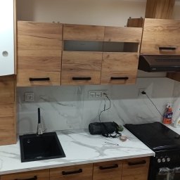 Kacper Starobrzyński - Nowoczesna kuchnia z drewnianymi szafkami, marmurowym blatem i czarnym zlewem. Widoczny bojler, okap i kuchenka. Uwagę zwraca wzór drewna na frontach szafek.