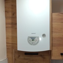 Kacper Starobrzyński - Biały piec gazowy Termet zamontowany na ścianie z drewnopodobnymi panelami, wkomponowany w zabudowę kuchenną z szafkami o podobnym wykończeniu.