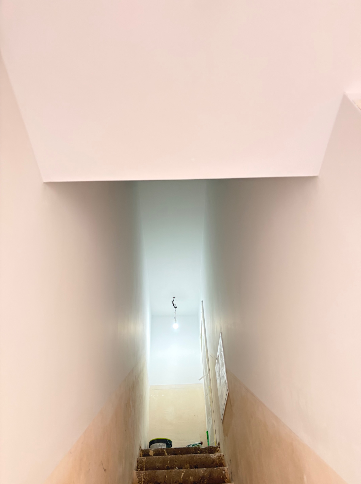 Klatka schodowa w trakcie malowania: górna część ścian i sufit pomalowane na biało, dolna część ścian w stanie surowym, widoczne schody i wiadro z farbą.