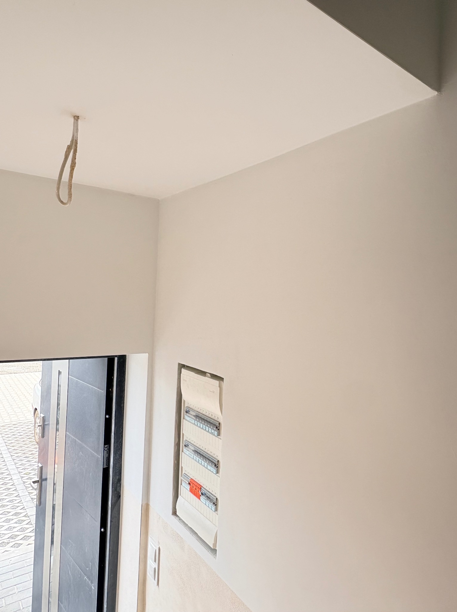 Świeżo pomalowane, jasne wnętrze z widoczną skrzynką elektryczną i wystającym kablem oświetleniowym. Drzwi wejściowe z widoczną kostką brukową na zewnątrz.