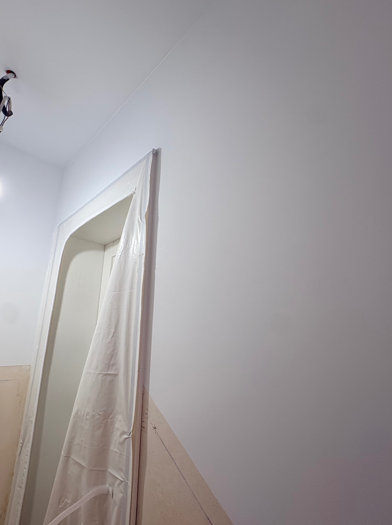 Przygotowanie do malowania: zabezpieczone folią drzwi w jasnym wnętrzu. Widoczne ślady ołówka na ścianie, sufit z instalacją elektryczną. Ujęcie z dołu.