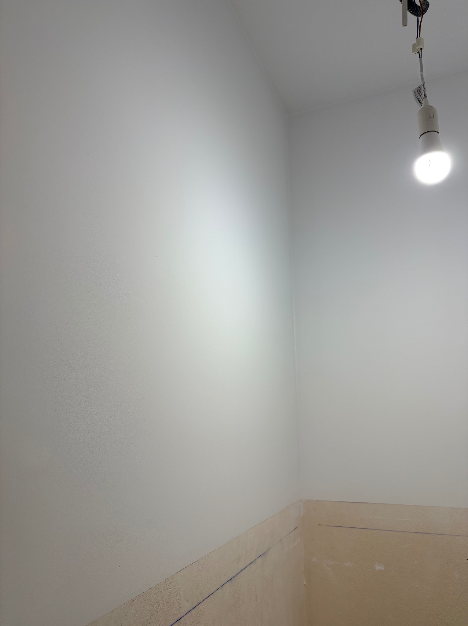 Świeżo pomalowane na biało ściany w rogu pokoju, widoczna żarówka na kablu i dolna część ściany przygotowana do dalszych prac wykończeniowych.