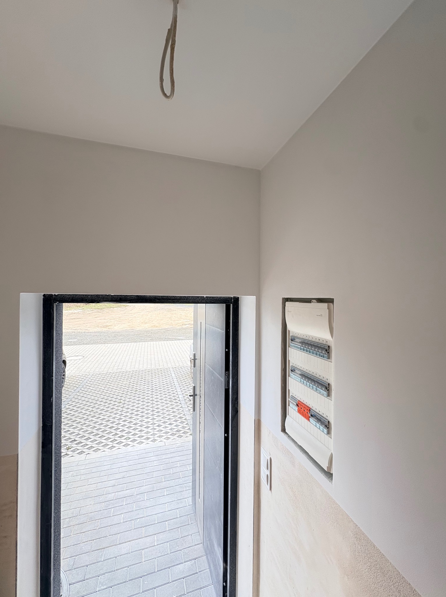 Wnętrze w trakcie wykańczania: świeżo pomalowane ściany w odcieniach bieli, otwarta stolarka drzwiowa z widokiem na kostkę brukową, skrzynka elektryczna w ścianie.