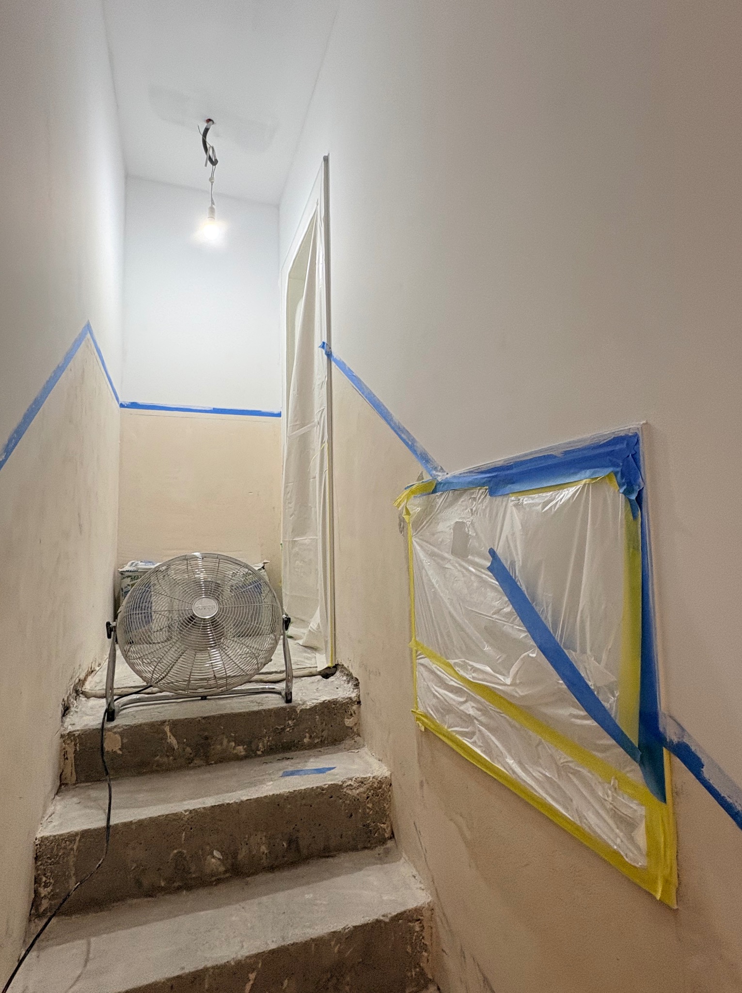 Klatka schodowa w trakcie malowania: ściany zabezpieczone taśmą malarską i folią, wentylator wspomaga schnięcie farby. Widoczne surowe schody i wisząca żarówka.
