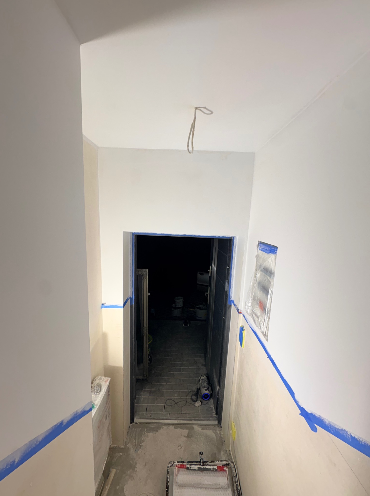 Korytarz w trakcie malowania: świeżo pomalowane ściany na biało, zabezpieczone taśmą malarską, widoczny wałek i puszki z farbą w tle. Oświetlenie sufitowe.