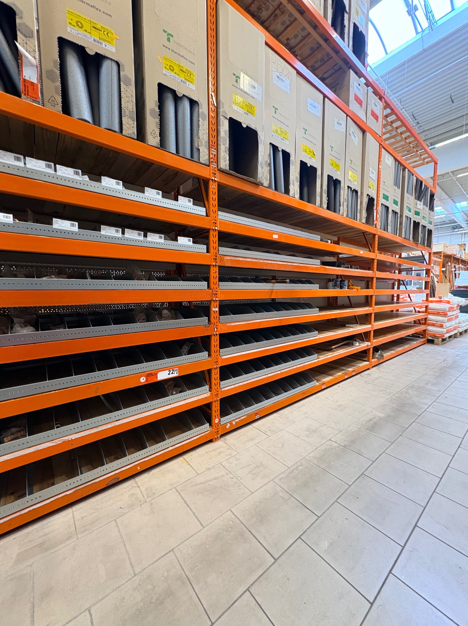 Pomarańczowy regał magazynowy z półkami, na których leżą kartony z izolacją termiczną. Widok z dołu, perspektywa zbieżna, jasne oświetlenie.