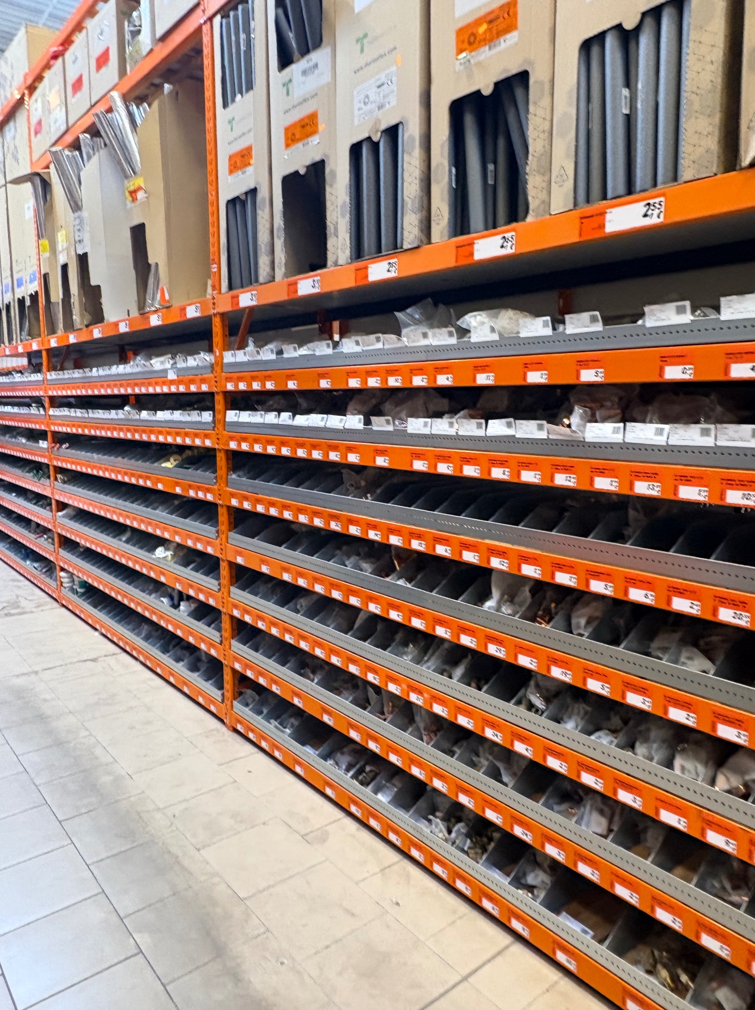 Półki magazynowe z materiałami budowlanymi, widok z perspektywy podłogi, pomarańczowe regały, szare podłogi, kartony z rurami na górnych półkach.