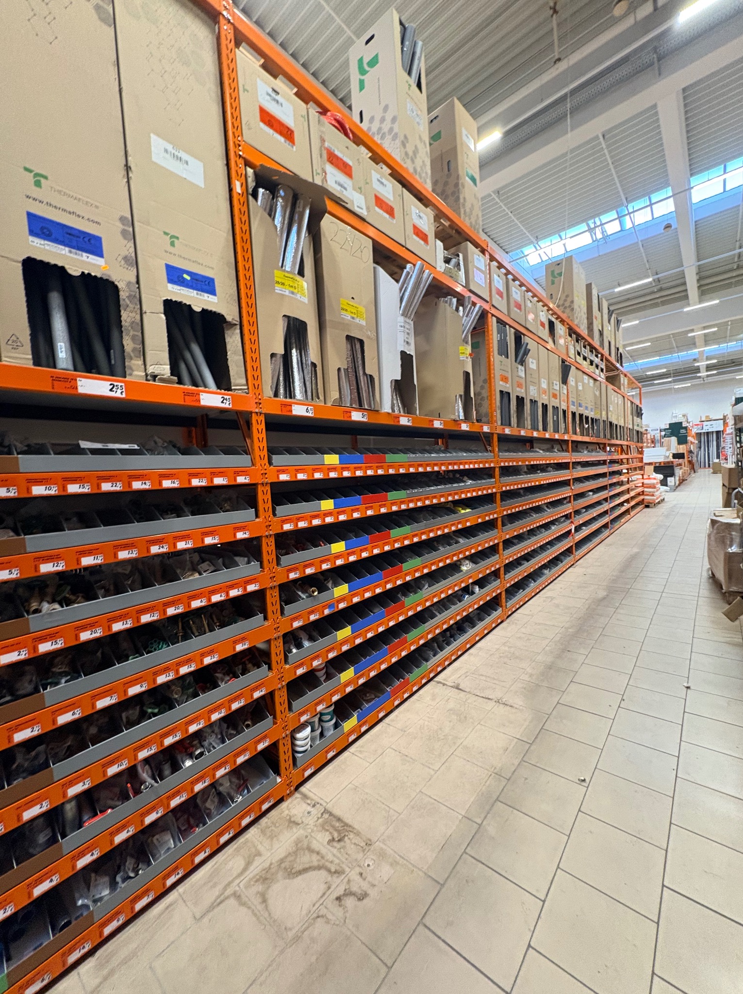 Długi regał magazynowy z rzędami półek wypełnionych materiałami instalacyjnymi i izolacyjnymi, widok z perspektywy alejki w sklepie budowlanym.