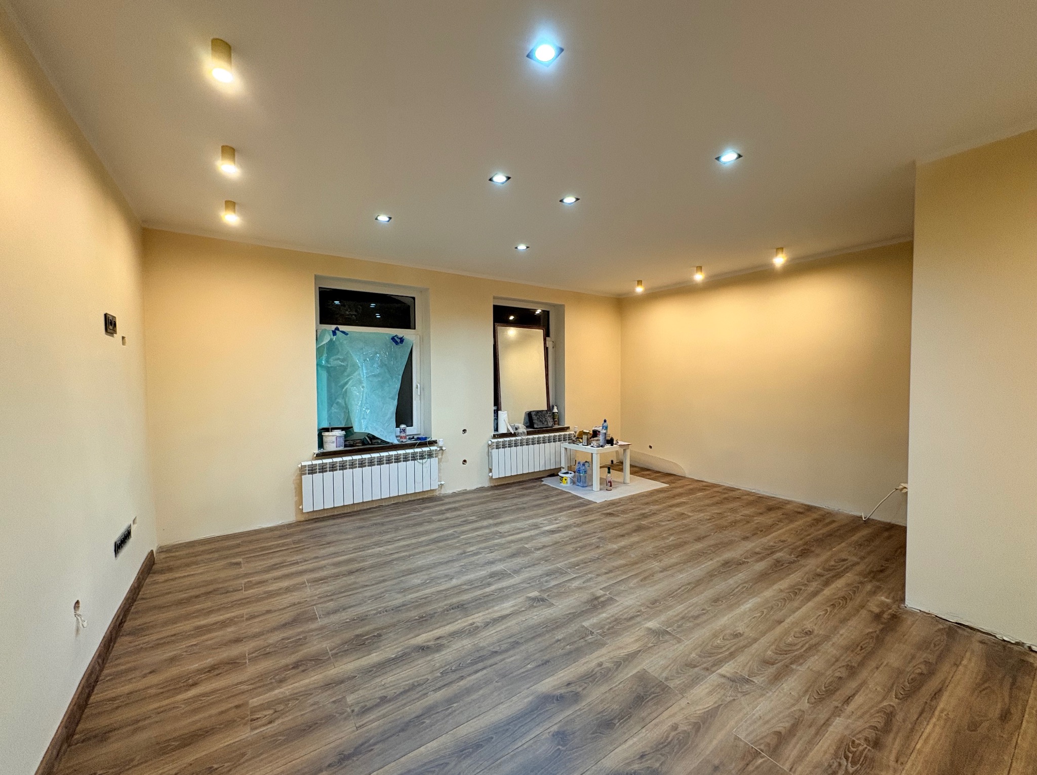 Jasne wnętrze po remoncie: brązowe panele podłogowe, beżowe ściany z punktowym oświetleniem. Okno zabezpieczone folią malarską, grzejnik. Widoczne ślady prac.