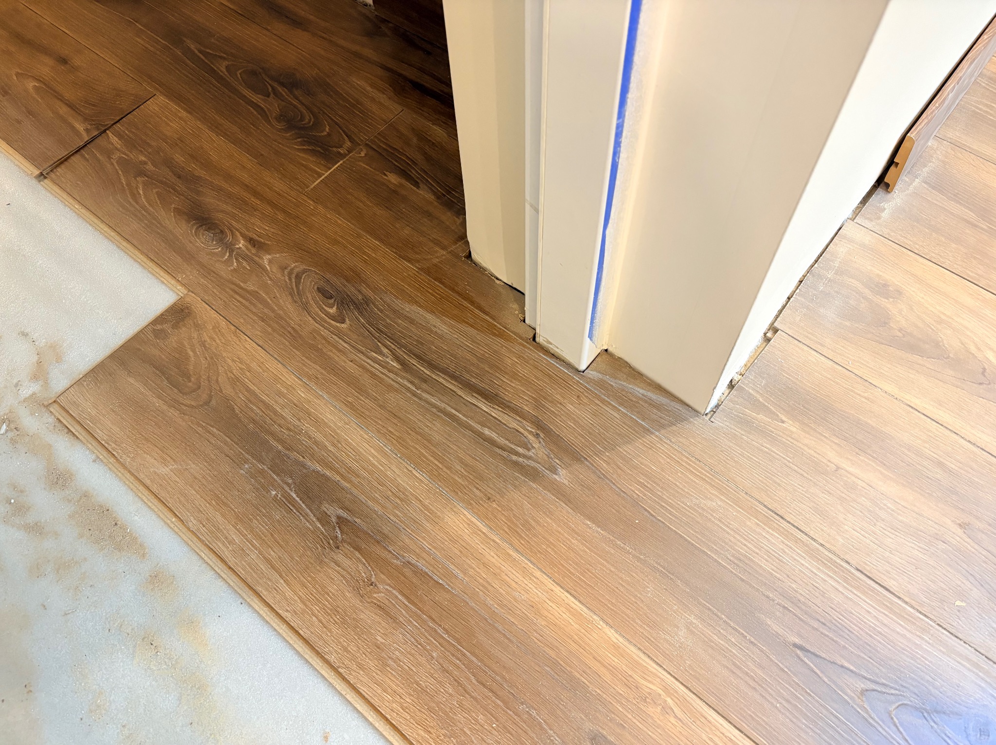 Układanie paneli podłogowych w odcieniach brązu przy futrynie drzwiowej, widoczna pianka podkładowa i szczelina dylatacyjna. Detal prac wykończeniowych.