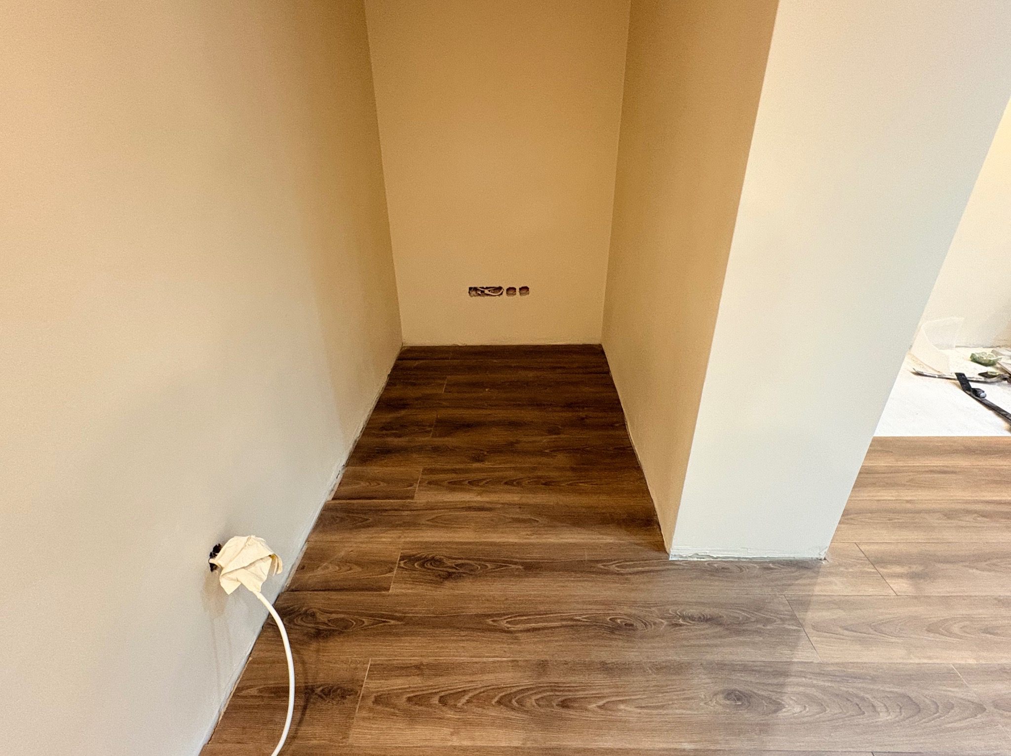 Wnęka z ułożonymi panelami podłogowymi w odcieniach brązu. Ściany pomalowane na beżowo, widoczne gniazdka elektryczne. Kabel wystający ze ściany zabezpieczony papierem.