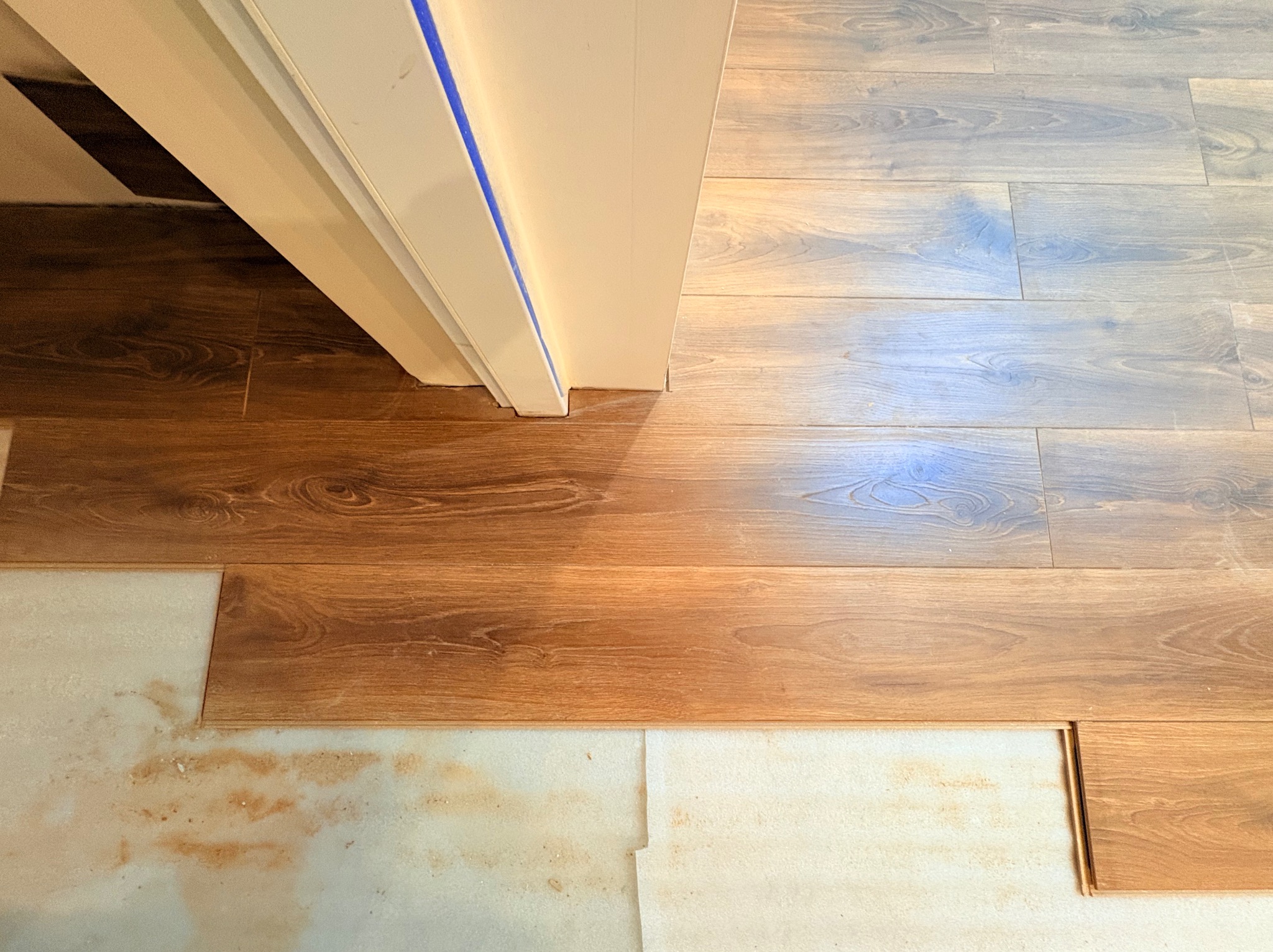 Układanie paneli podłogowych w odcieniach brązu i szarości, widoczne podłoże izolacyjne i fragment ościeżnicy z niebieską taśmą malarską.