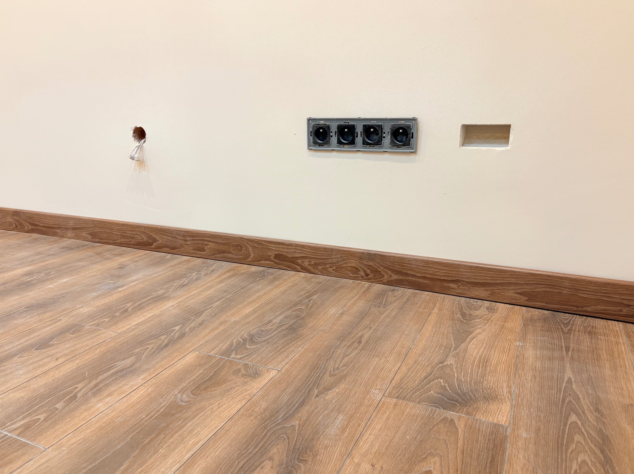 Ściana z gniazdkami w trakcie montażu i fragmentem drewnopodobnej podłogi z listwą przypodłogową. Widoczne otwory na kable i puszki elektryczne.