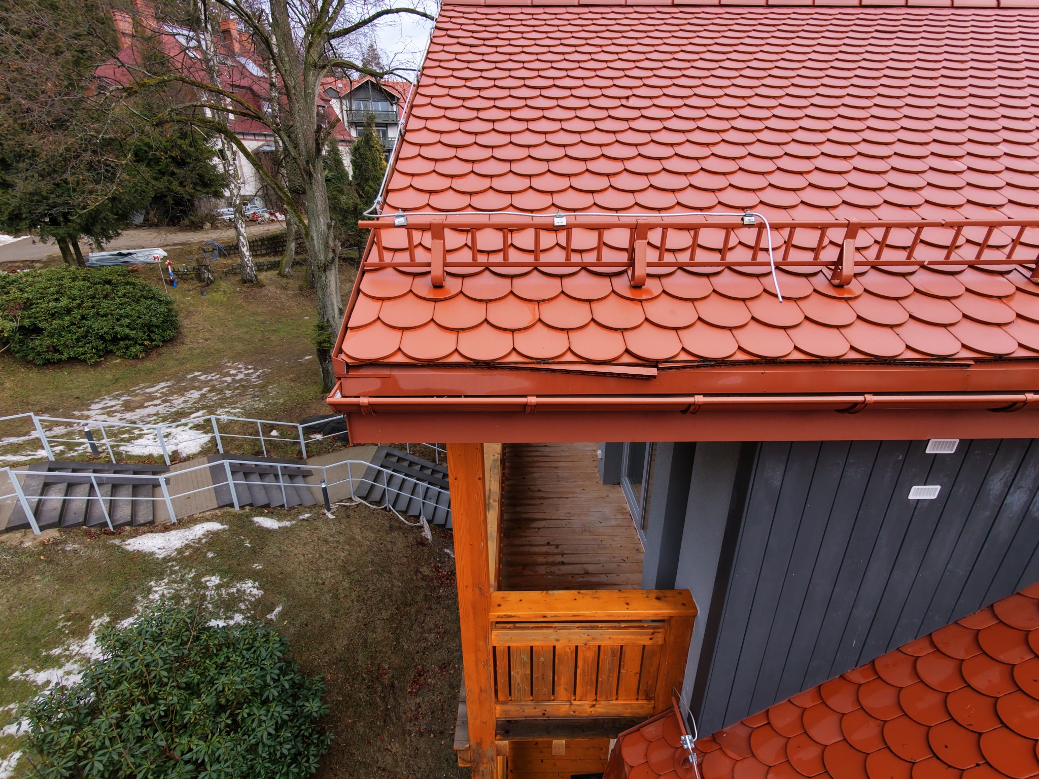 Widok z góry na fragment dachu z czerwoną dachówką karpiówką, drewniany balkon i schody terenowe. W tle budynek i drzewa.