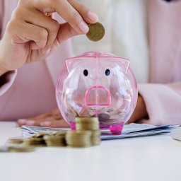 Phinance - Ręka wkłada monetę do przezroczystej świnki skarbonki, obok stos banknotów i monet. Koncepcja oszczędzania, inwestycji i planowania finansowego.