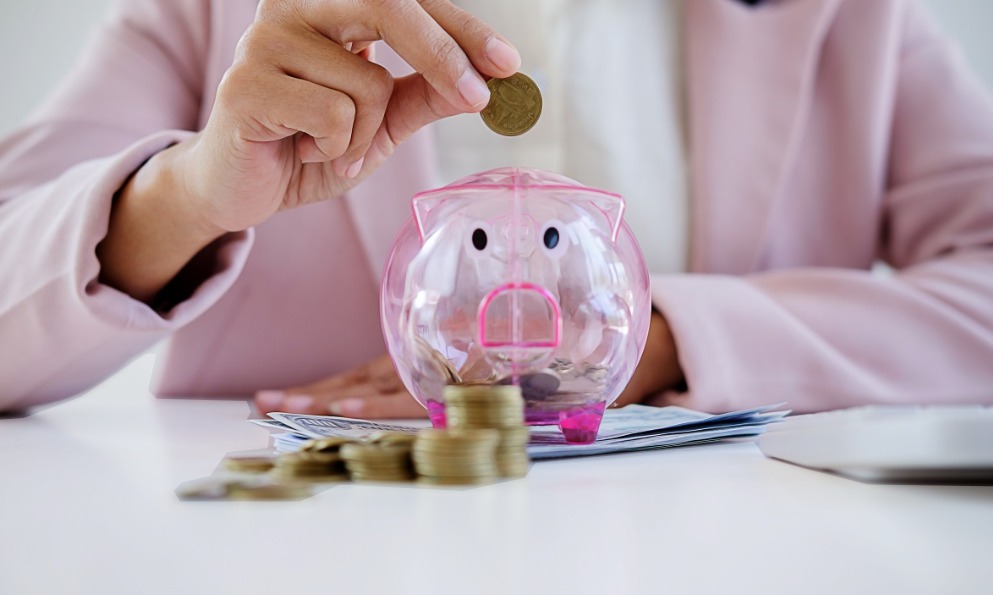 Ręka wkłada monetę do przezroczystej świnki skarbonki, obok stos banknotów i monet. Koncepcja oszczędzania, inwestycji i planowania finansowego.