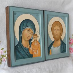 Anastasiia Dolhu - Dwie ręcznie malowane ikony religijne w drewnianych, błękitnych ramkach, przedstawiające Matkę Boską z Dzieciątkiem oraz Jezusa Chrystusa, ustawione na białym obrusie z haftem.