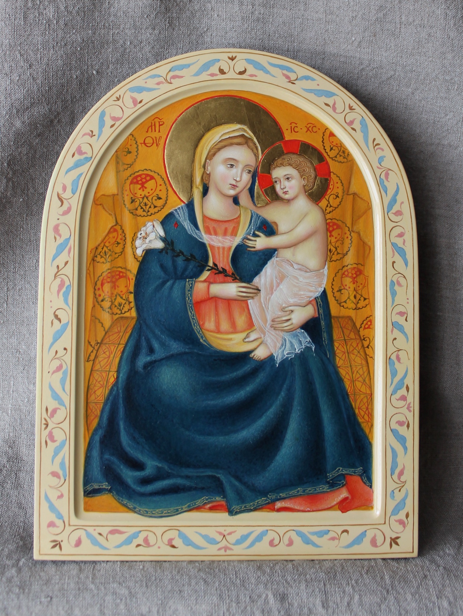 Ręcznie malowana ikona religijna przedstawiająca Matkę Boską z Dzieciątkiem Jezus, w bogato zdobionej ramie z motywami roślinnymi na szarym tle.