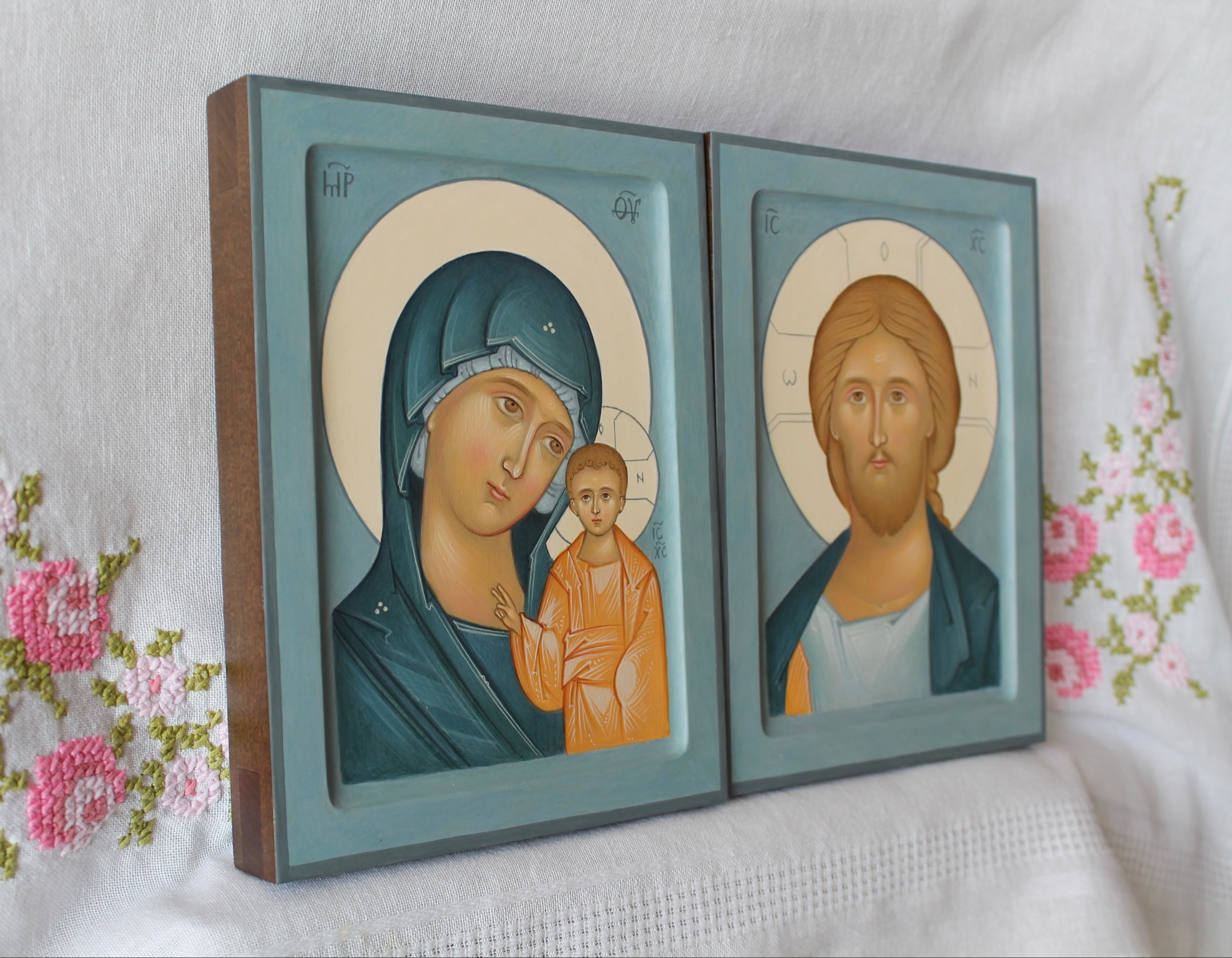 Dwie ręcznie malowane ikony religijne w drewnianych, błękitnych ramkach, przedstawiające Matkę Boską z Dzieciątkiem oraz Jezusa Chrystusa, ustawione na białym obrusie z haftem.