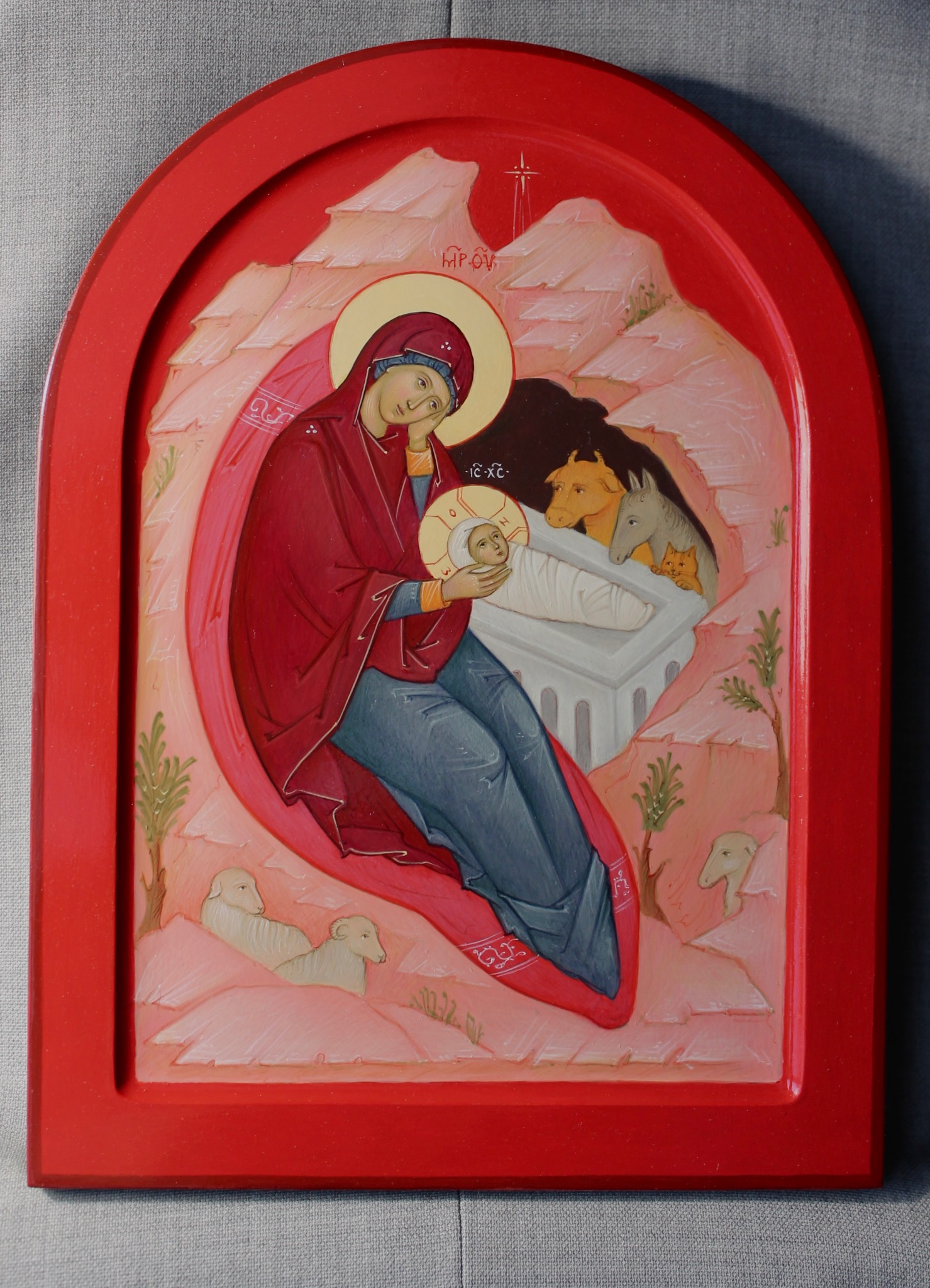 Ręcznie malowana ikona religijna przedstawiająca Matkę Boską z Dzieciątkiem Jezus w czerwonej ramie, z motywami zwierząt i krajobrazu. Delikatne, pastelowe barwy.