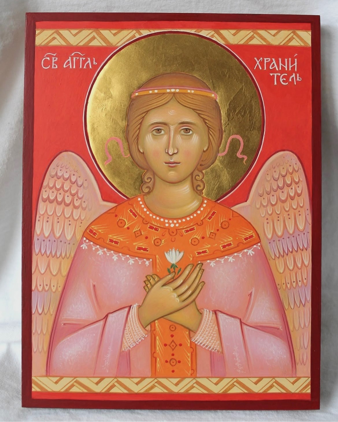 Ręcznie malowana ikona religijna przedstawiająca anioła z aureolą, trzymającego biały kwiat na czerwonym tle z ornamentami. Widoczne skrzydła i inskrypcje w języku cerkiewnosłowiańskim.