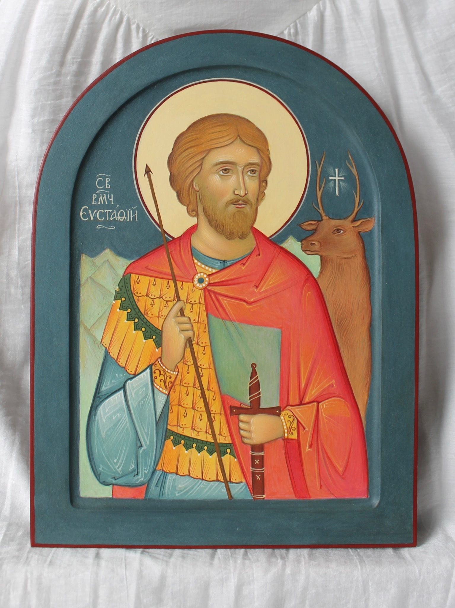 Ręcznie malowana ikona religijna przedstawiająca św. Eustachego z jeleniem, na drewnianej desce w niebieskiej ramie, z widocznymi detalami pędzla i fakturą farby.