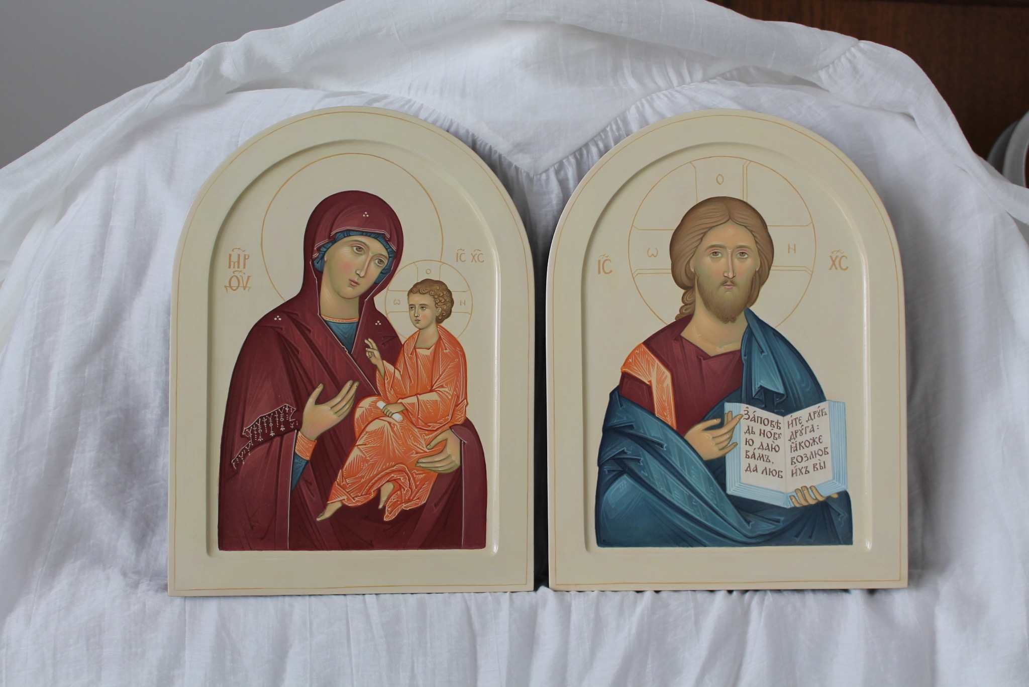 Dwie ręcznie malowane ikony religijne na kremowym tle, przedstawiające Matkę Boską z Dzieciątkiem i Jezusa Chrystusa z księgą, ukazane w zbliżeniu.
