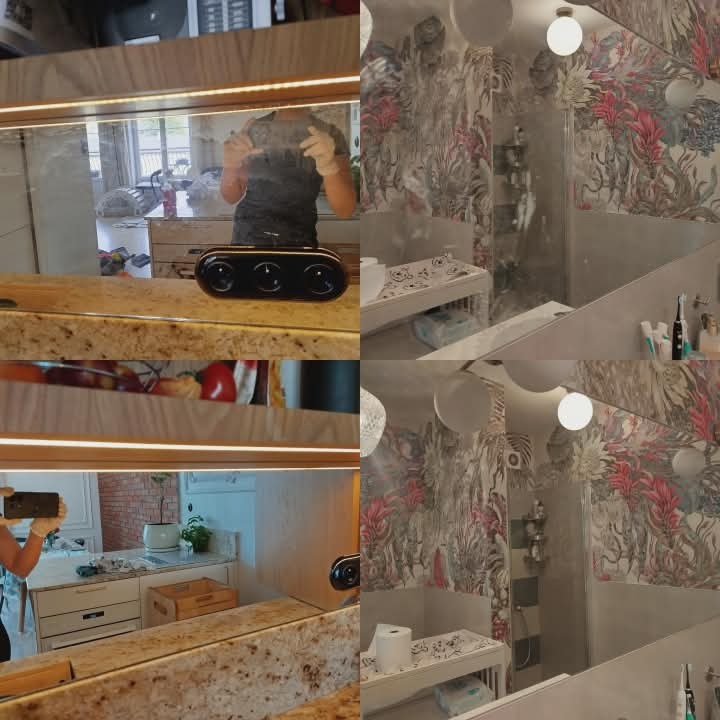 Odbicie osoby w rękawicach czyszczącej lustro w łazience z wzorzystą tapetą. Widoczne akcesoria łazienkowe i oświetlenie LED. Ujęcie z bliska, skupione na detalu.