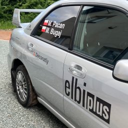 ELBI plus Arkadiusz Pacan - Srebrny samochód z naklejkami firmowymi 'Decoratly' i 'elbiplus' na karoserii oraz naklejkami z imionami w oknach, zaparkowany na żwirowym podłożu.