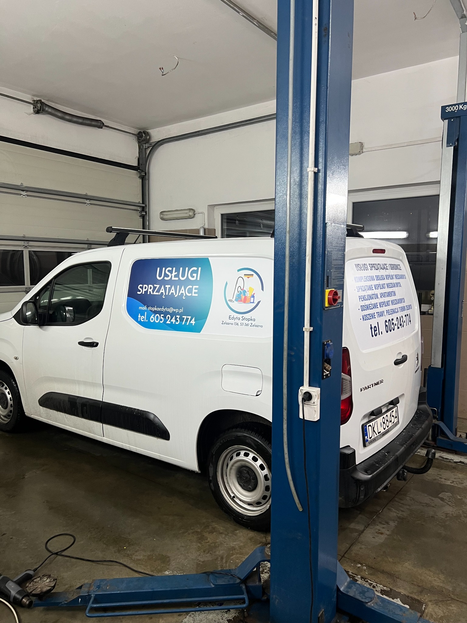 Biały samochód dostawczy z logo firmy sprzątającej w warsztacie samochodowym. Widoczne reklamy usług sprzątania na karoserii i numer telefonu.