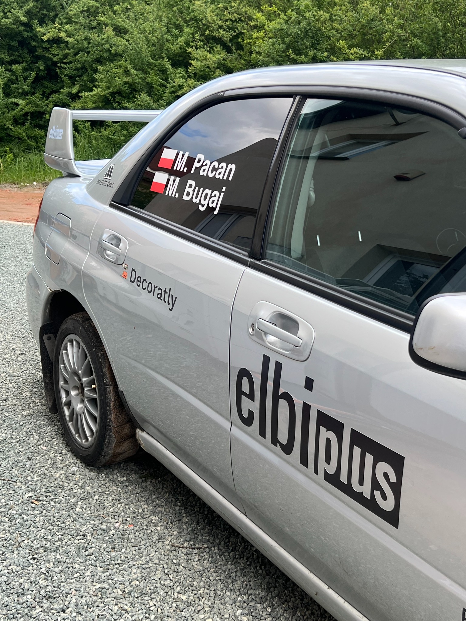 Srebrny samochód z naklejkami firmowymi 'Decoratly' i 'elbiplus' na karoserii oraz naklejkami z imionami w oknach, zaparkowany na żwirowym podłożu.