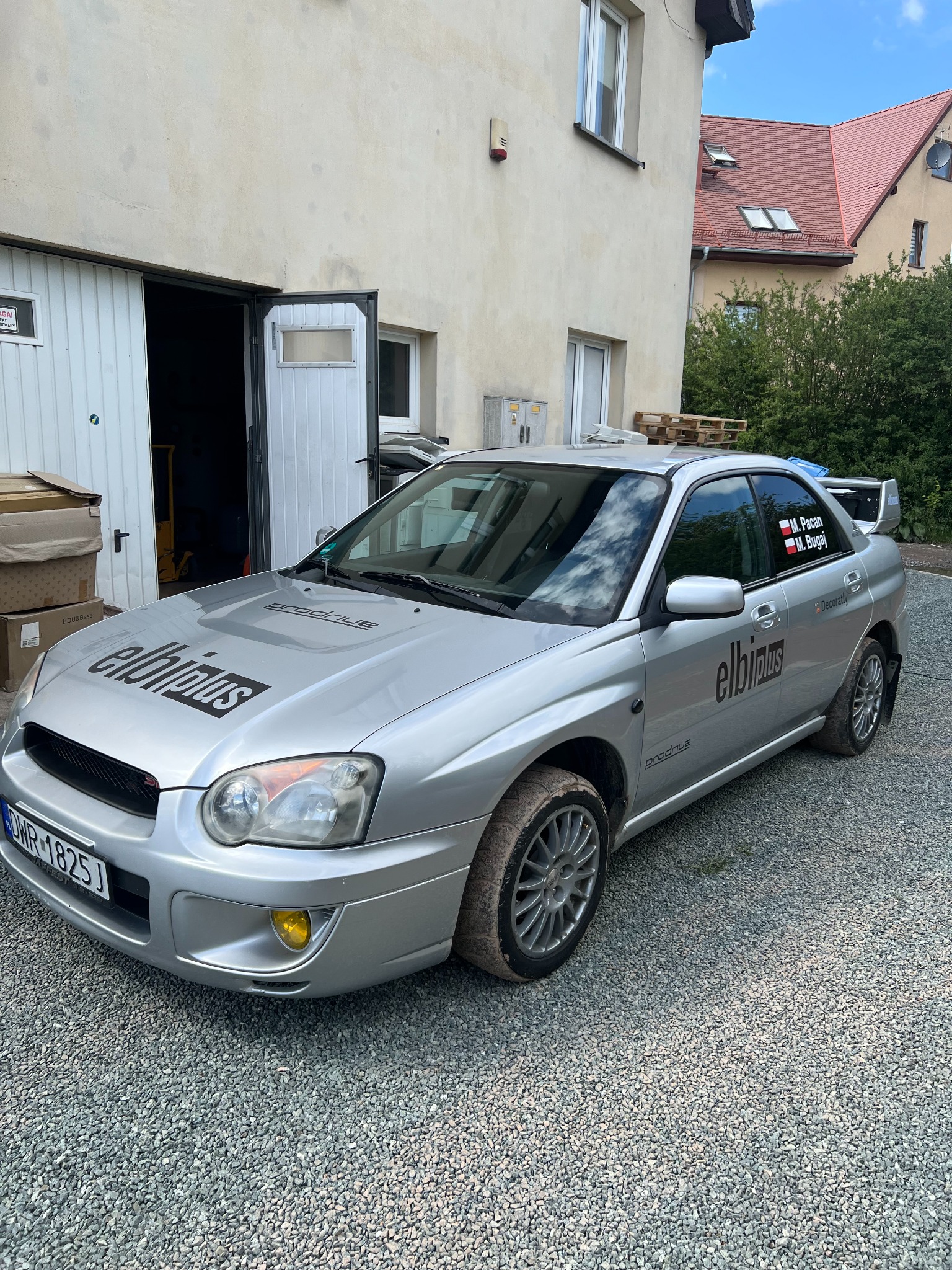 Srebrny samochód Subaru Impreza z naklejonym logo 'elbipius' na masce i boku, zaparkowany na żwirowym podjeździe przed budynkiem z otwartymi drzwiami garażowymi.