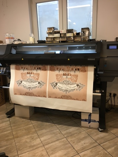 Drukarka wielkoformatowa w trakcie druku dwóch identycznych plakatów z ozdobnym motywem i tekstem w języku polskim. Widoczne kartony i okno w tle.