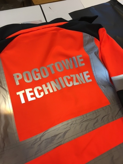 Pomarańczowa kurtka robocza z odblaskowymi pasami i srebrnym napisem 'POGOTOWIE TECHNICZNE' na plecach, widok z tyłu, leżąca na białym podłożu.