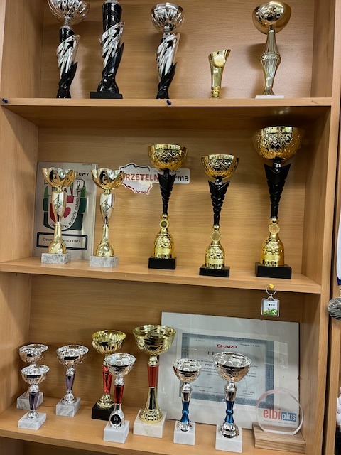 Półka z trofeami w Kłodzku: różnorodne puchary złote, srebrne i czarne, certyfikat Sharp, breloczek z logiem. Trofea sportowe na drewnianej półce.