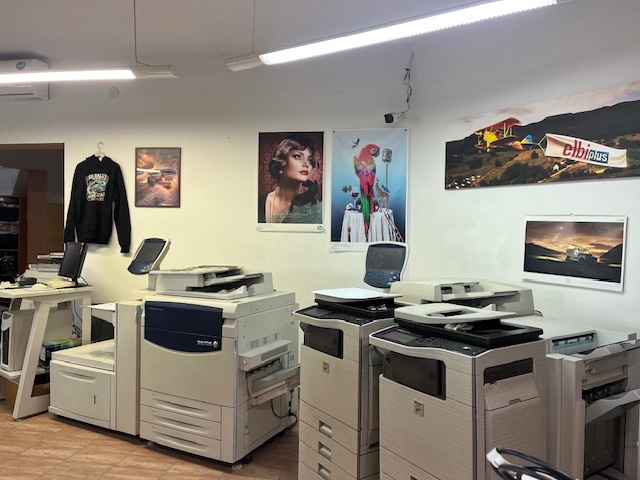 Wnętrze drukarni z kilkoma urządzeniami drukującymi, plakatami na ścianach i bluzą z kapturem wiszącą obok. Widoczne biurko z komputerem i inne akcesoria biurowe.